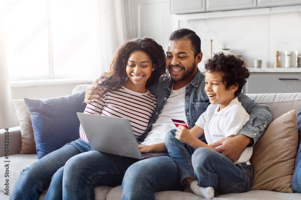 African American man, woman, and child are seated on a sofa. They are all looking at a laptop screen, engaged in an activity. The room is well-lit and cozy, creating a warm atmosphere. African American man, woman, and child are seated on a sofa. They are all looking at a laptop screen, engaged in an activity. The room is well-lit and cozy, creating a warm atmosphere.