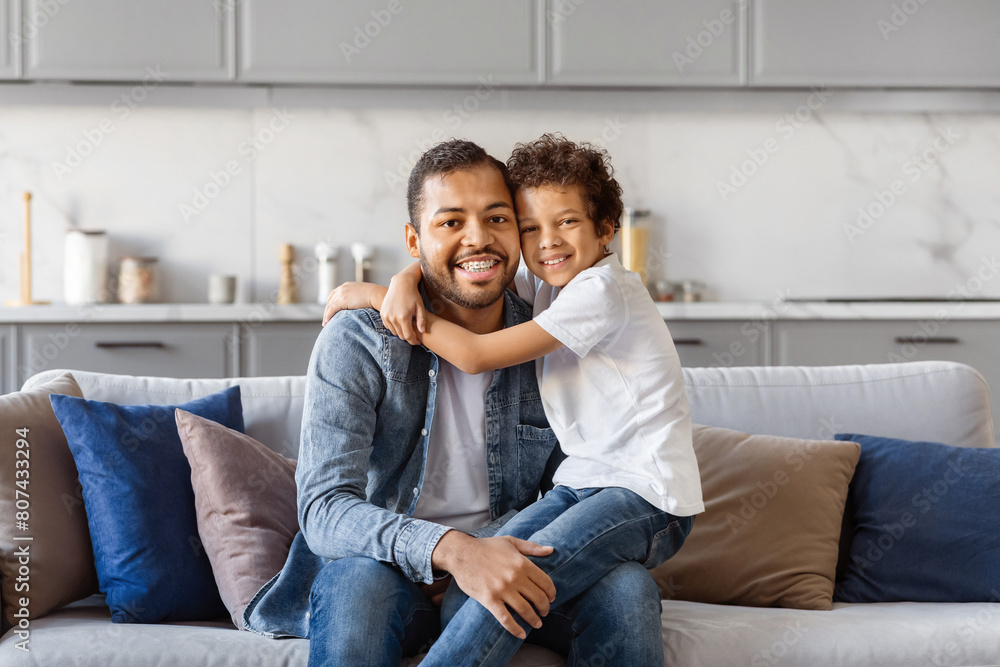 African American man is sitting on top of a couch next to a little boy. The father appears relaxed, while the son looks curious. They seem to be engaged in a conversation or activity together. African American man is sitting on top of a couch next to a little boy. The father appears relaxed, while the son looks curious. They seem to be engaged in a conversation or activity together.