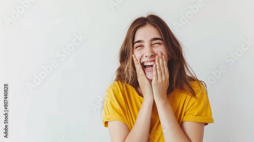Young woman in yellow t-shirt laughing with hands on face against white background. photo