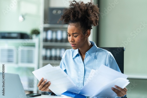 Analyzing business charts A4, a young African American woman in a blue formal shirt and afro brown hair works as a Market Research Analyst in a modern office. photo