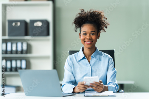 Performing calculations with a calculator, a young African American woman in a blue formal shirt with afro brown hair works as a Market Research Analyst in a modern office. photo