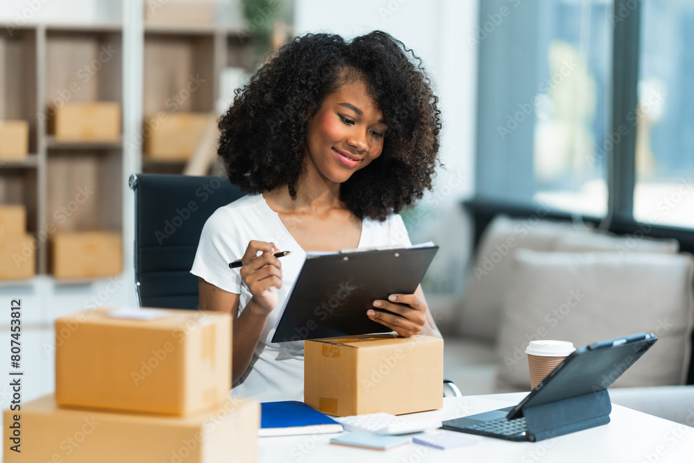 A young African American woman with afro brown hair works in a modern office, managing her online clothing store and live stream platform, sme box packages. A young African American woman with afro brown hair works in a modern office, managing her online clothing store and live stream platform, sme box packages.