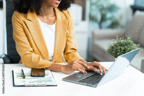 Performing calculations with a calculator, a young African American woman in yellow formal suit with afro brown hair works as a Market Research Analyst in a modern office. photo