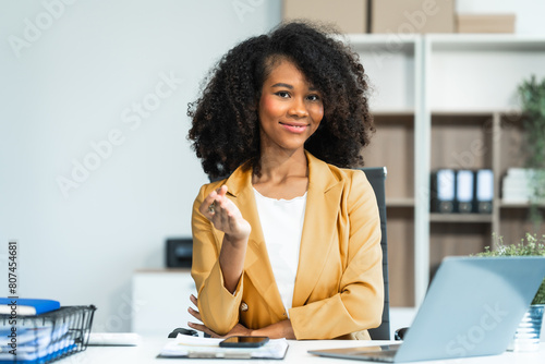 Performing calculations with a calculator, a young African American woman in yellow formal suit with afro brown hair works as a Market Research Analyst in a modern office. photo