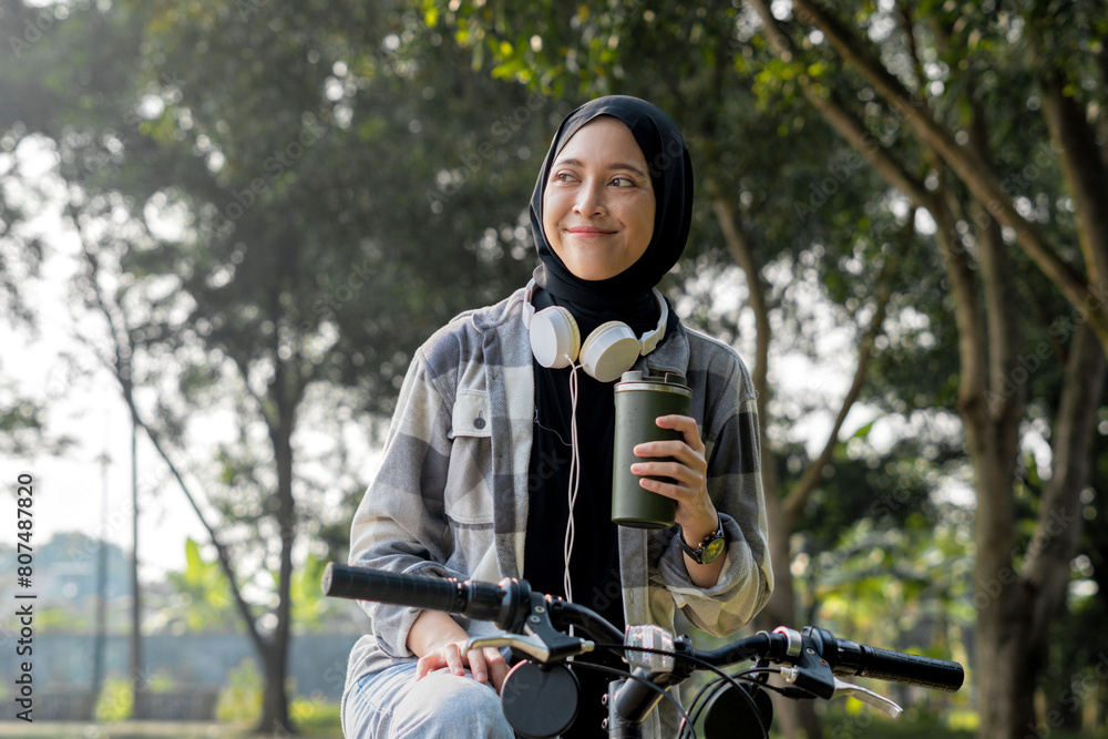 Fototapeta premium Young cheerful hijab woman, wearing casual flannel riding bicycle bike on sidewalk in the city park enjoying being outdoors, look aside or ahead. People active urban healthy lifestyle cycling concept.