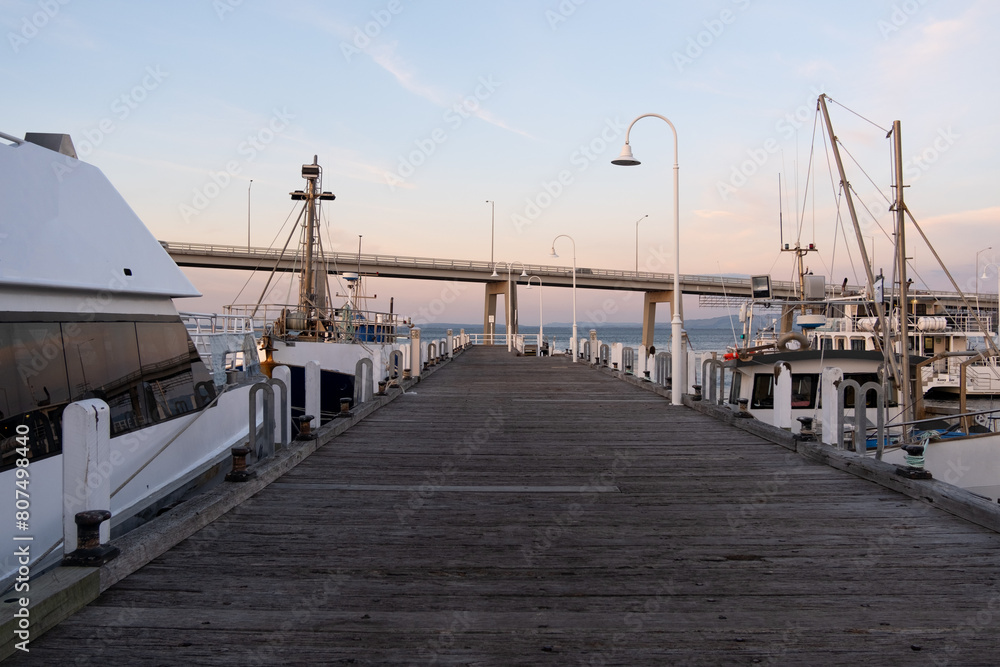 Fototapeta premium Wooden boardwalk at San Remo Jetty with ships berthed and Phillip Island bridge in the distance during sunset. Background texture of a timber pier at the shore with copy space.
