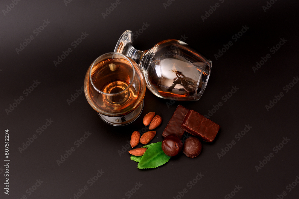 Two glasses of cognac on a black background, next to an almond, mint leaves and chocolates. Two glasses of cognac on a black background, next to an almond, mint leaves and chocolates.