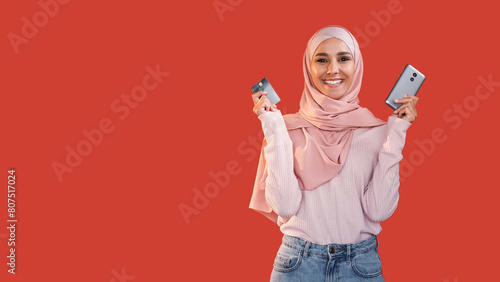 Mobile banking. Online shopping. Satisfied cheerful happy woman in hijab showing credit card phone isolated on red empty space background. © golubovy