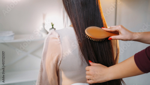 Professional hairstylist. Salon treatment. Woman hands combing model lady beautiful smooth long brunette hair with wooden brush in light interior. © golubovy