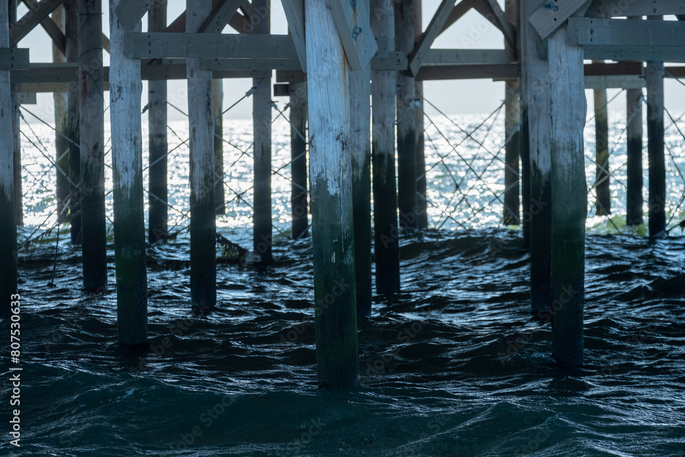 This image offers a serene yet powerful view beneath a pier, where sunlight dances on the sea's surface beyond the sturdy, barnacle-clad pillars, casting deep shadows and highlighting the textures of This image offers a serene yet powerful view beneath a pier, where sunlight dances on the sea's surface beyond the sturdy, barnacle-clad pillars, casting deep shadows and highlighting the textures of