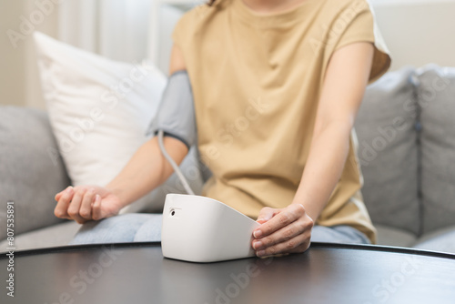 Hypotension problem, close up hand of  young woman check pulse, measure high blood pressure heart rate, holding digital pressure monitor, making self on arm, cuff on sofa at home. Healthcare medical. © KMPZZZ