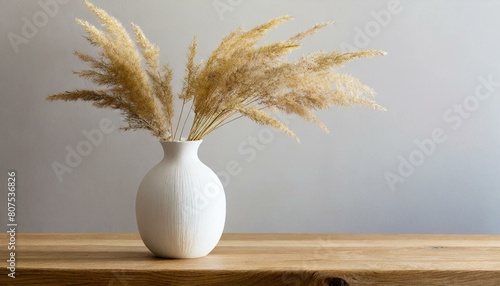 Modern white ceramic vase with dry grass on wooden table. Scandinavian interior. Empty white wall,