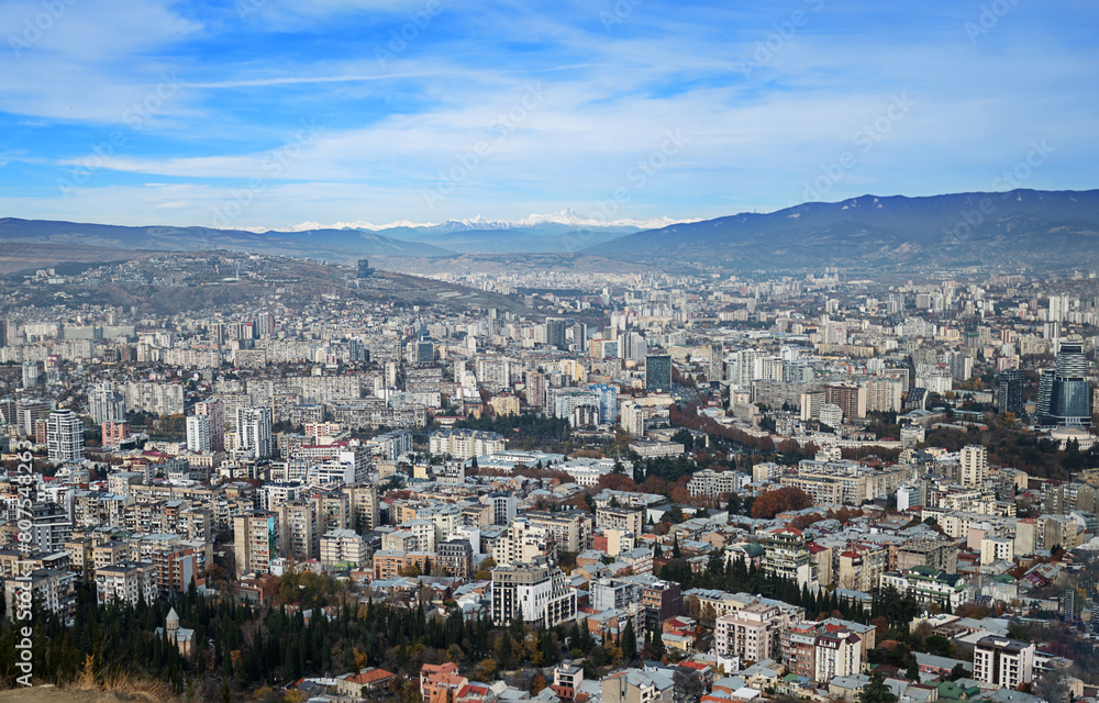sprawling urban landscape of Tbilisi, Georgia, under a clear blue sky view showcases diverse architectural styles densely packed residential areas stretching towards the distant mountains
