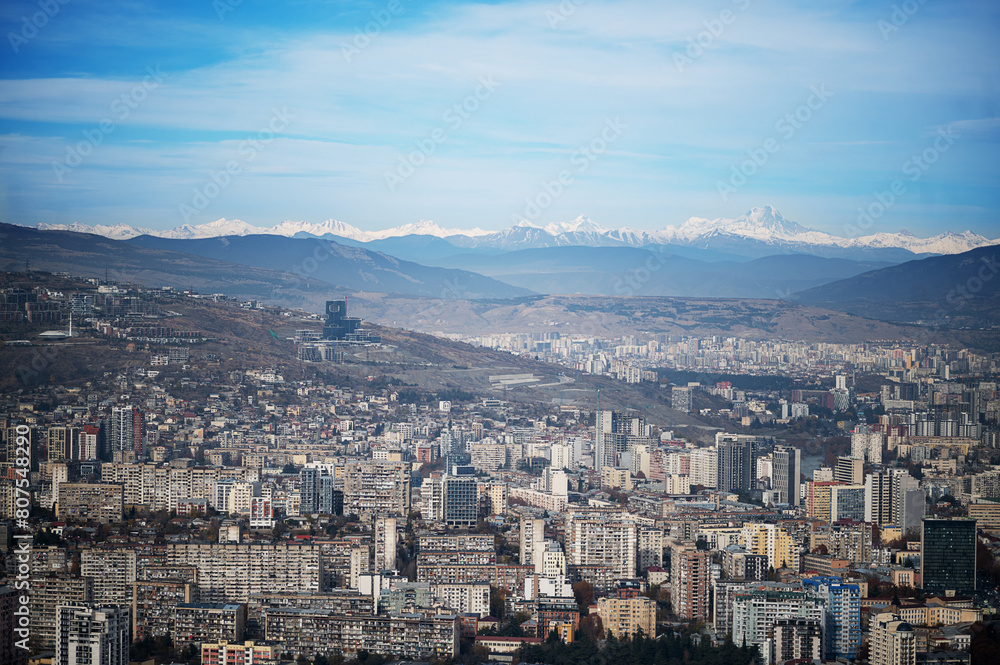 Obraz premium expansive aerial view of Tbilisi, Georgia, showcasing a dense urban landscape with various buildings under a clear blue sky, bordered by mountains in the distance