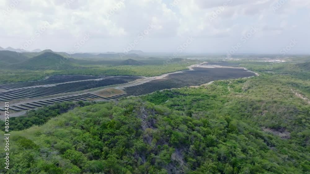 Ascending drone shot of large solar panel power station park with green scenery during cloudy day. Rainforest mountains of Dominican Republic. Wide shot. Ascending drone shot of large solar panel power station park with green scenery during cloudy day. Rainforest mountains of Dominican Republic. Wide shot.