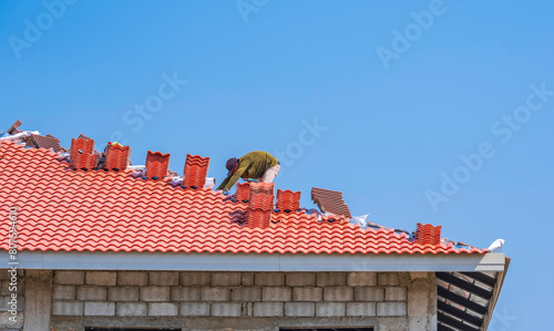Construction worker is laying aluminum foil coated insulation and orange roof tiles on hip roof structure of modern house building against blue clear sky background © Prapat
