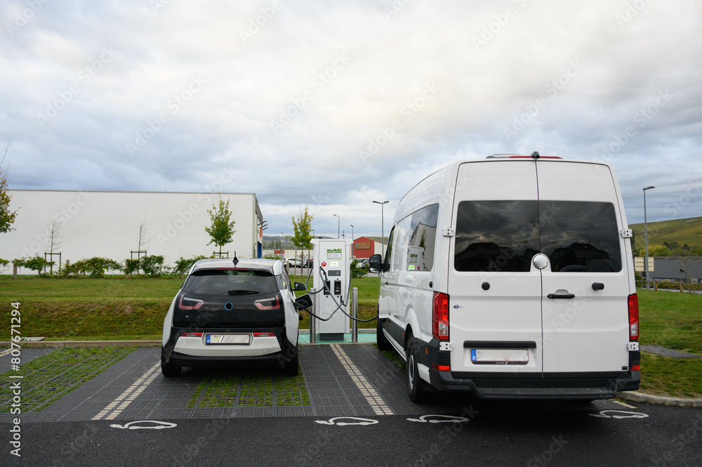 An electric car and a an electric van are charging at a photovoltaic station in a parking. Electric car pictograms on the ground.