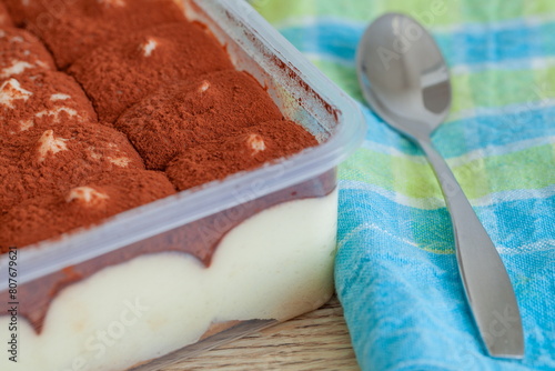 Tiramisu cake in plastic box and spoon on wooden background