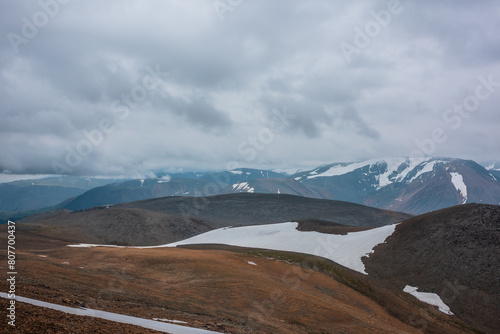 Dark overcast landscape with high pass, wide glacier on stony hill slope and snowy mountain range silhouette in far away in rainy low clouds. Large mountains with snow under gray sky in bad weather.