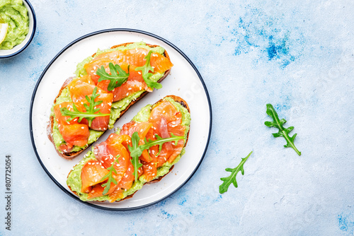 Avocado toast with salmon on rye bread with guacamole sauce, arugula and sesame seeds on plate, blue table background, top view © 5ph