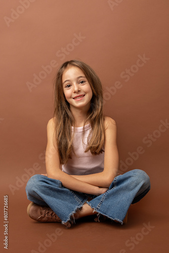 Funny Girl in casual clothes sitting on floor and looking at camera against brown background. Studio photography