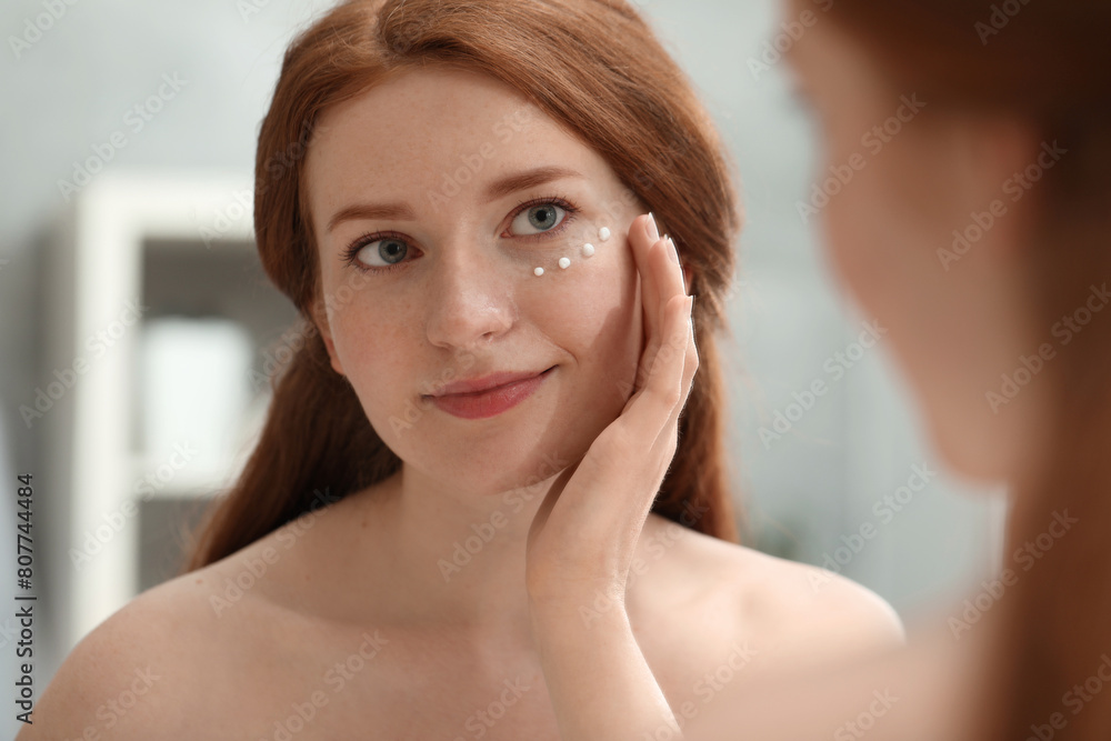 Beautiful woman with freckles applying cream onto her face near mirror in bathroom Beautiful woman with freckles applying cream onto her face near mirror in bathroom