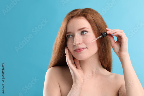 Beautiful woman with freckles applying cosmetic serum onto her face against light blue background. Space for text