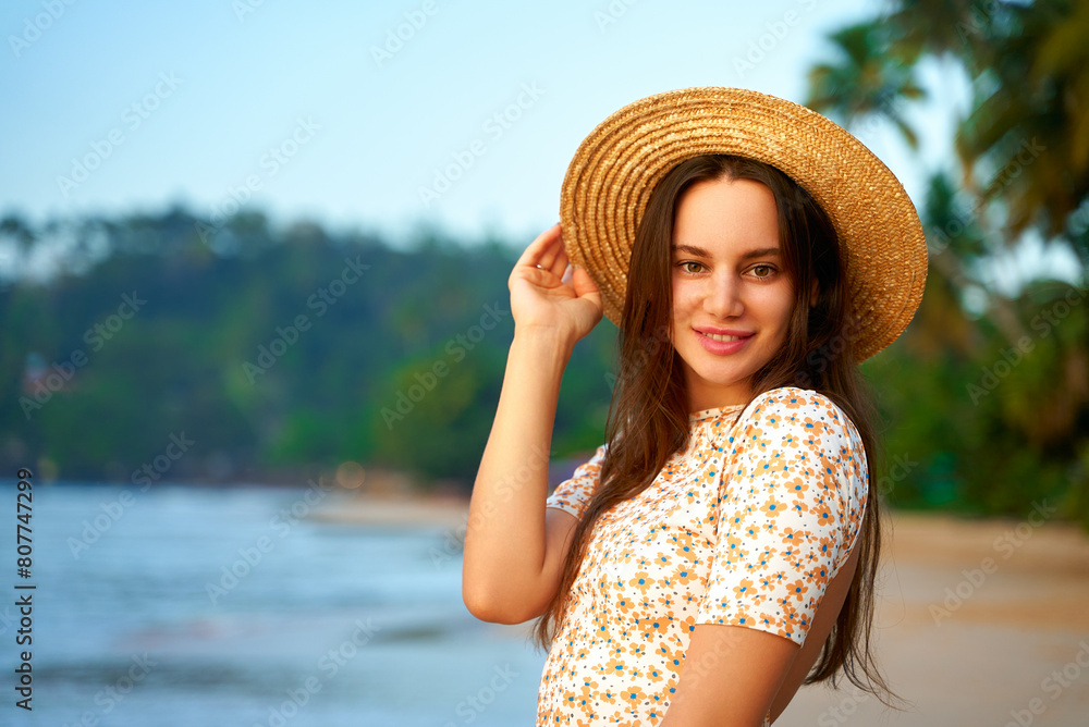 Smiling woman in floral dress holds straw hat on tropical beach at sunset. Happy female enjoys summer evening by sea, palm trees in background. Traveler poses during golden hour on island coast.