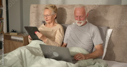  Senior couple using a digital tablet and a laptop together in their bed