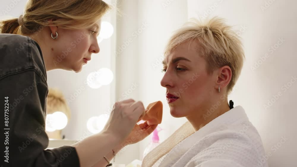 Waist up of makeup artist touching up face of Caucasian elegant blonde woman sitting in front of backstage mirror with bulbs and preparing for studio portrait photoshoot Waist up of makeup artist touching up face of Caucasian elegant blonde woman sitting in front of backstage mirror with bulbs and preparing for studio portrait photoshoot