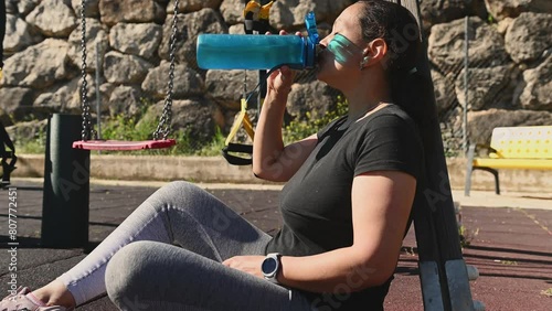 Happy smiling fit woman in activewear drinking water after fitness workout with suspension straps outdoors. Sportswoman holding bottle of water, relaxing after sports training on urban sportsground photo