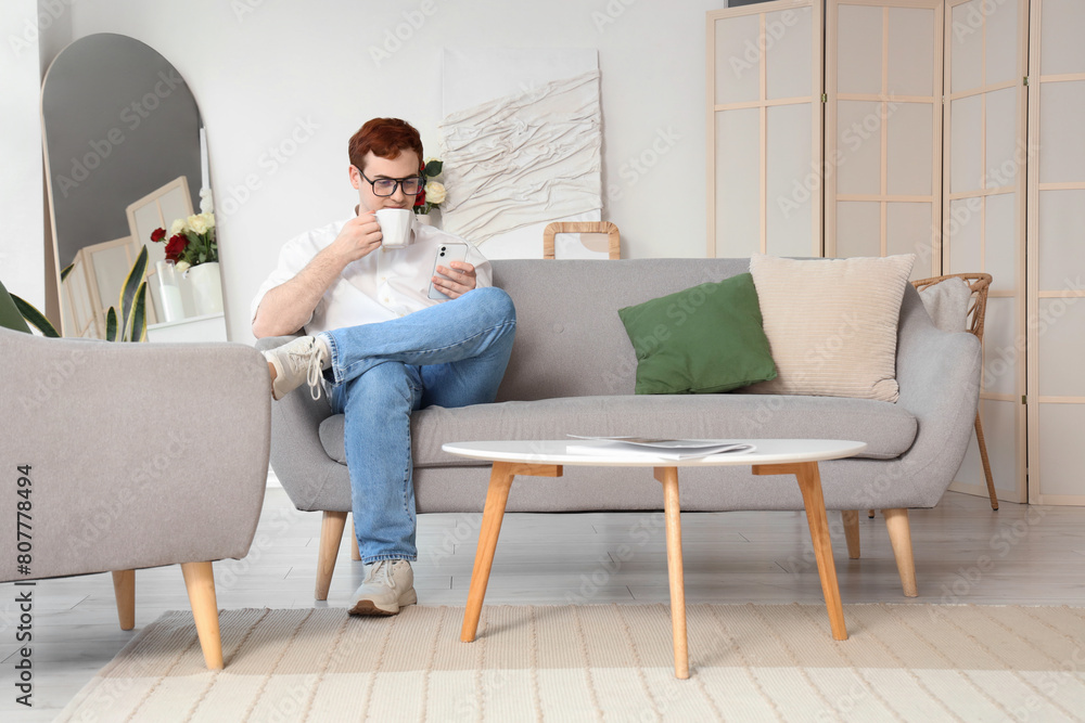 Obraz premium Young man in eyeglasses with cup of tea and mobile phone sitting on grey sofa at home