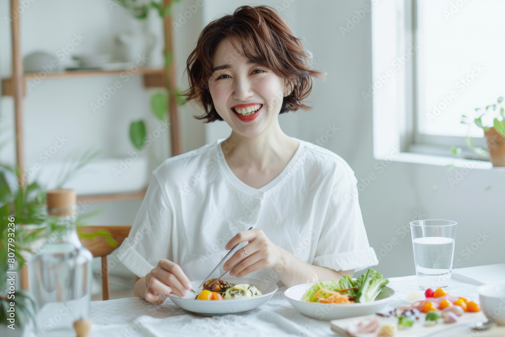 A Japanese woman in her thirties is smiling while eating, with short hair and wearing white. She is sitting at the dining table of an all-white modern minimalist kitchen