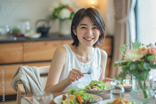 A Japanese woman in her thirties is smiling while eating  with short hair and wearing white. She is sitting at the dining table of an all-white modern minimalist kitchen