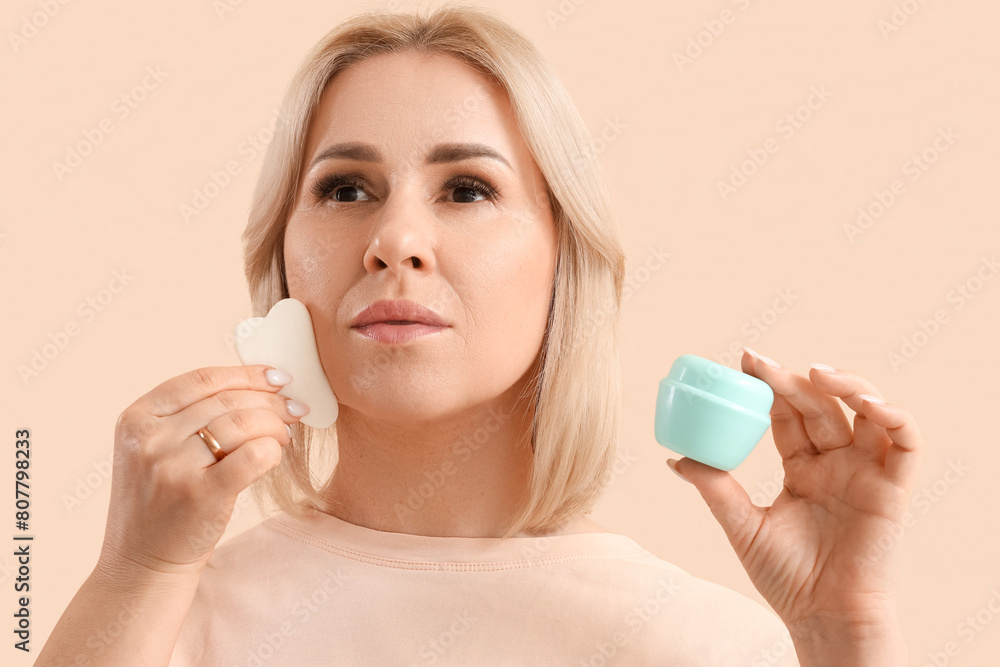 Beautiful mature woman with jar of facial cream and guasha tool on beige background Beautiful mature woman with jar of facial cream and guasha tool on beige background