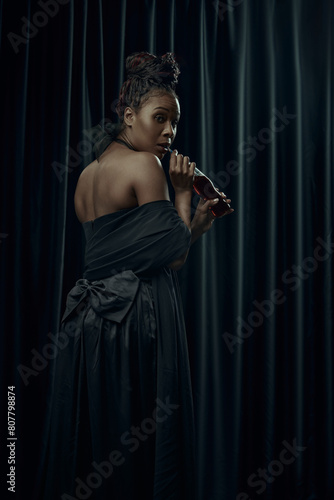 Young African-American woman, dressed as medieval person in classical black dress drinking soda against dark curtain backdrop. Concept of comparison of eras, modernity and history.