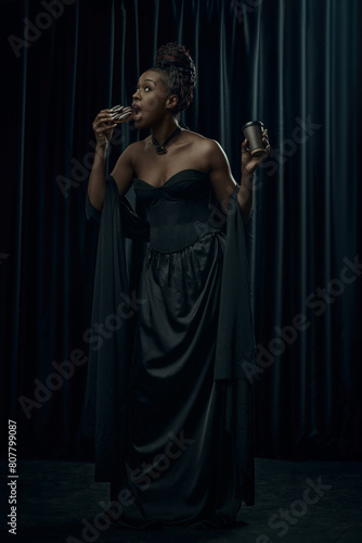 African-American woman dressed as medieval person eating and drinking modern food against dark curtain backdrop. Concept of food and drink, comparison of eras, modernity and history.