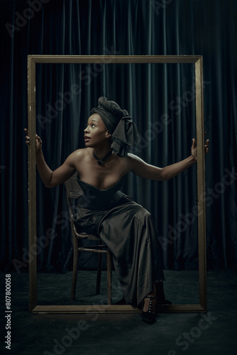 African-American woman posing inside large, ornate frame, dressed in sophisticated black gown with regal headwrap against dark curtain backdrop. Concept of comparison of eras, modernity and history.