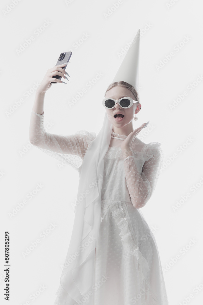 Young woman in white costume with conical hat, and modern sunglasses, looks as medieval person, taking selfie against white studio background. Concept of history, renaissance art, comparison of eras. Young woman in white costume with conical hat, and modern sunglasses, looks as medieval person, taking selfie against white studio background. Concept of history, renaissance art, comparison of eras.