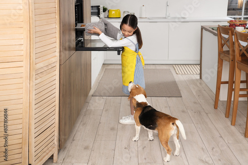 Cute little Asian girl with Beagle dog taking cake from oven in kitchen