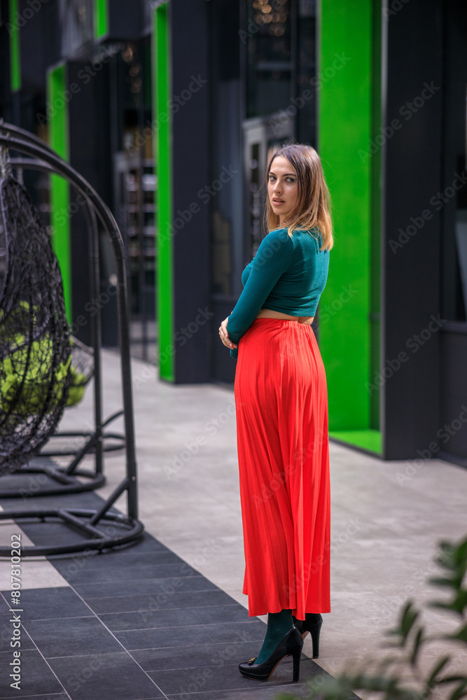 A young woman in bright clothes, a red skirt and a green longsleeve is standing in a business center. High-heeled shoes. A young woman in bright clothes, a red skirt and a green longsleeve is standing in a business center. High-heeled shoes.