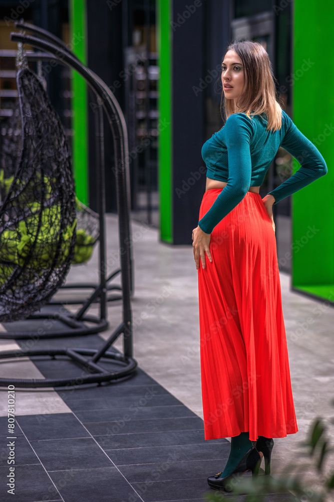 A young woman in bright clothes, a red skirt and a green longsleeve is standing in a business center. High-heeled shoes. A young woman in bright clothes, a red skirt and a green longsleeve is standing in a business center. High-heeled shoes.