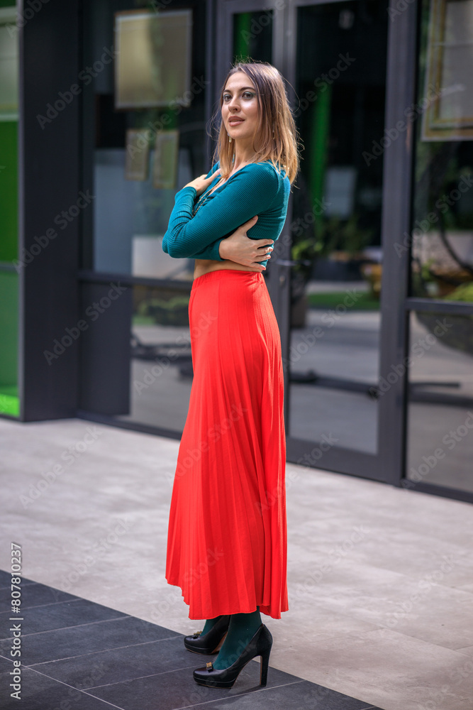 A young woman in bright clothes, a red skirt and a green longsleeve is standing in a business center. High-heeled shoes. A young woman in bright clothes, a red skirt and a green longsleeve is standing in a business center. High-heeled shoes.