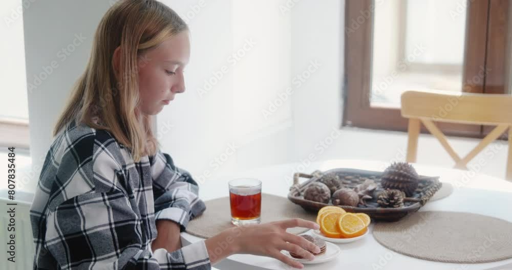 Serious girl eating crackers while drinking tea. Female teenager is sitting at table in room. She is enjoying snacks at home during sunny day. Serious girl eating crackers while drinking tea. Female teenager is sitting at table in room. She is enjoying snacks at home during sunny day.