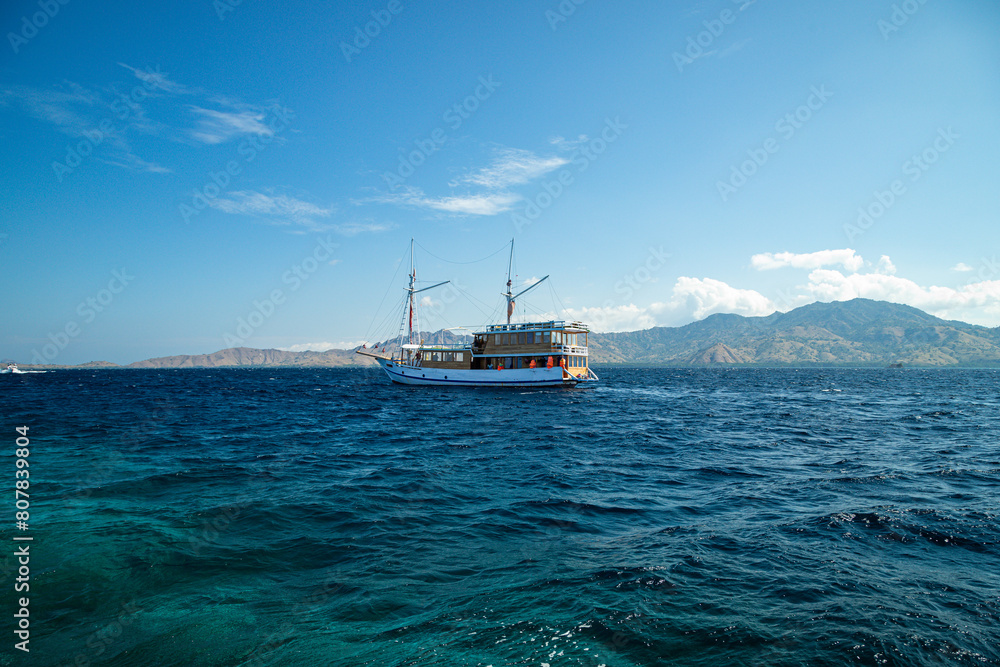 Sailing through the azure sea, a pinisi ship navigates amidst lush green hills, with the clear blue sky as its backdrop.