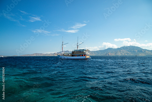 Sailing through the azure sea, a pinisi ship navigates amidst lush green hills, with the clear blue sky as its backdrop.