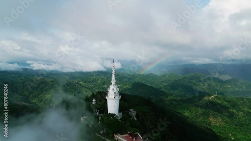 Aerial drone circles Ambuluwawa Tower in Sri Lanka, reveals rainbow above lush green mountains, symbolizes harmony, pilgrimage site surrounded by clouds, spiritual getaway spot in serene highlands. photo