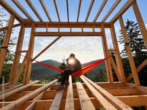 Man on construction site of wooden-framed house sits in hammock against backdrop of mountains and sunrise, sipping coffee. The concept pertains to modern ecological construction.