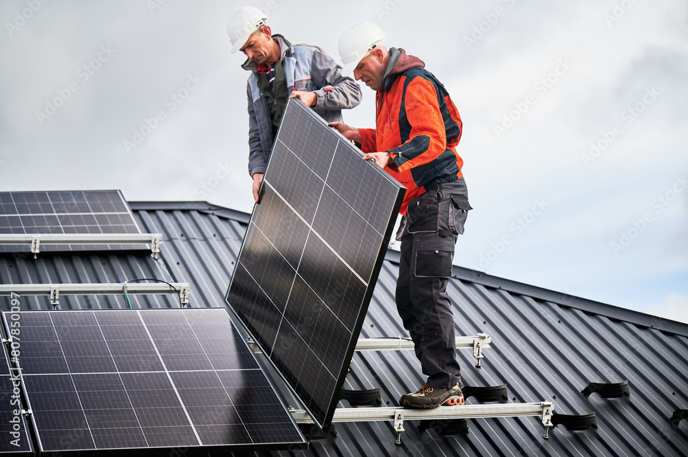 Mounters building photovoltaic solar module station on roof of house. Men electricians in helmets installing solar panel system outdoors. Concept of alternative and renewable energy. Mounters building photovoltaic solar module station on roof of house. Men electricians in helmets installing solar panel system outdoors. Concept of alternative and renewable energy.
