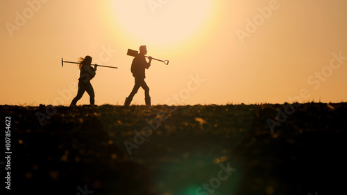 Silhouettes of two farmers walking with working tools across a field against the backdrop of a beautiful sunset.